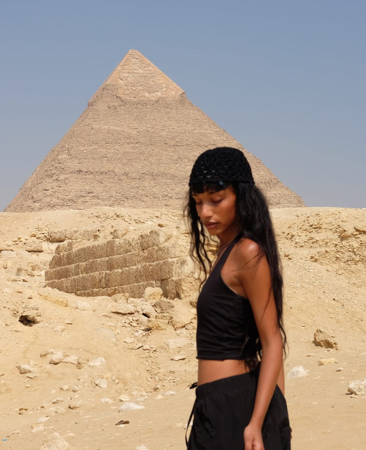 Woman in a black crochet hat standing in front of a pyramid in the desert